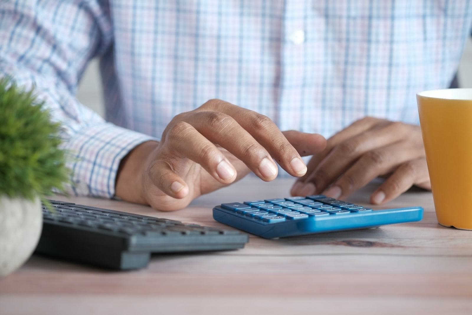 A person working out a figure on a blue desk calculator beside a keyboard and a mug of coffee
