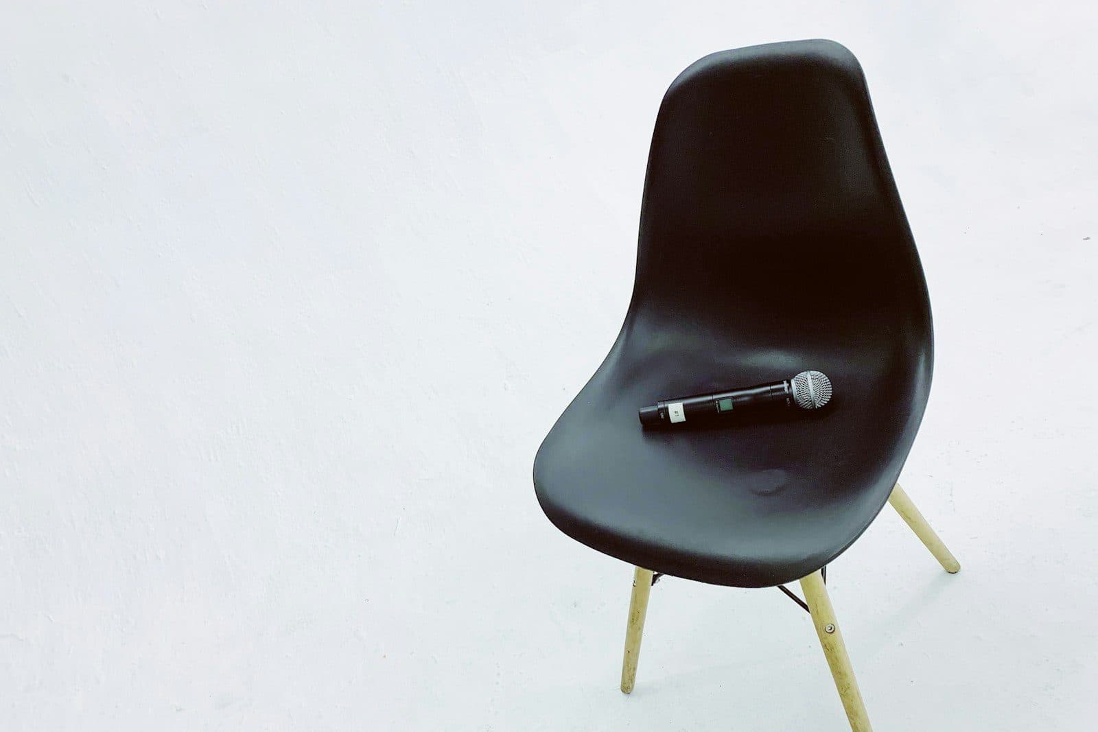 A single black chair against a white wall with a wireless microphone lying on the seat