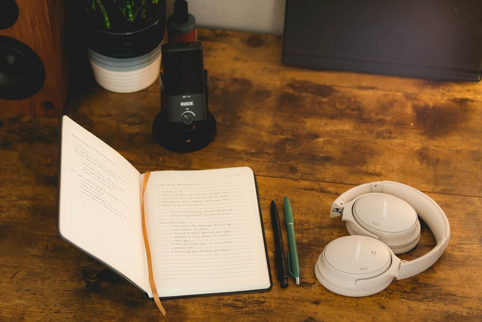 An open handwritten notebook, two pens and headphones on a wooden desk beside a desktop microphone