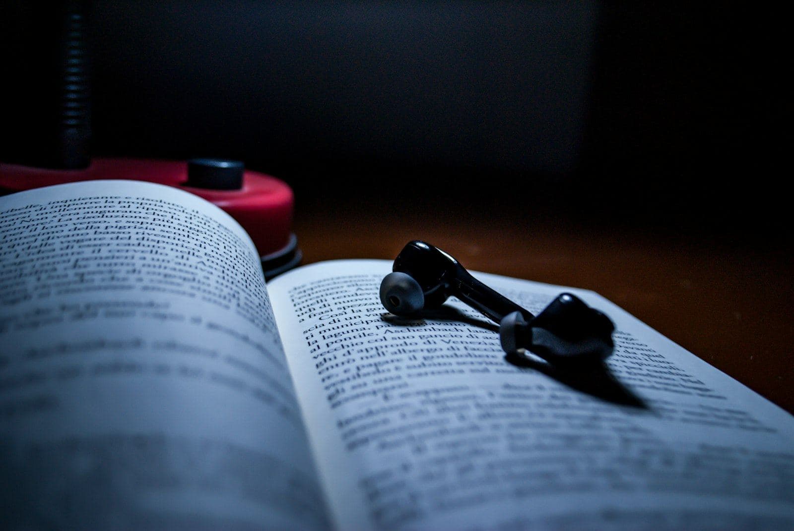 A pair of black earbuds resting on the open pages of a paperback book on a dark desk