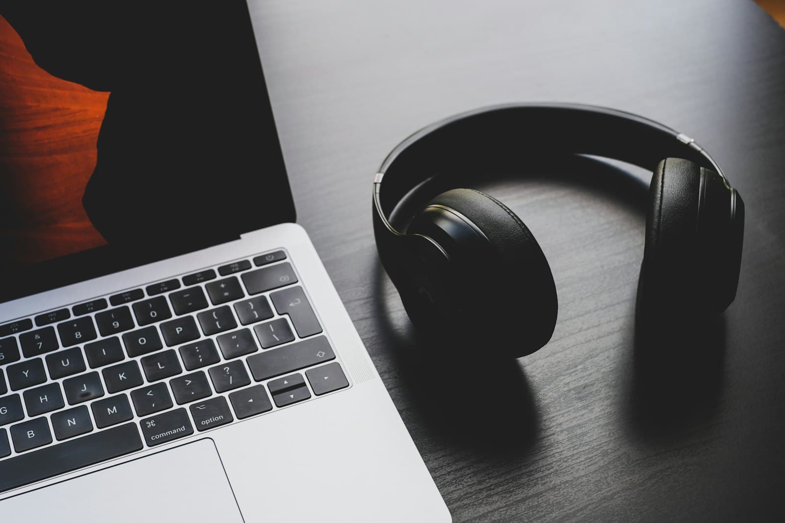 Black over-ear headphones beside an open silver laptop on a dark wooden desk