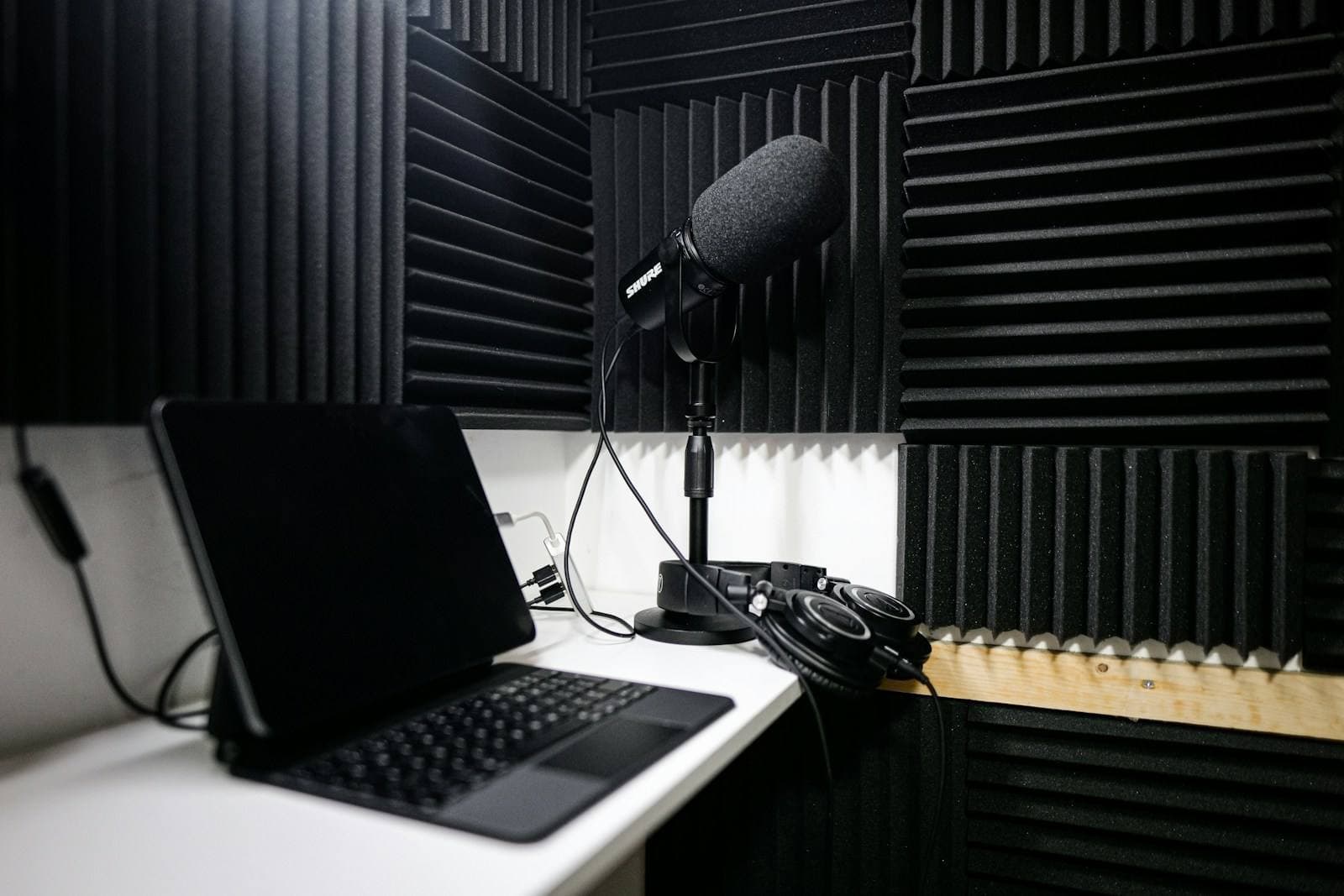 A small recording booth lined with acoustic foam, with a microphone on a stand, a tablet and headphones on the desk