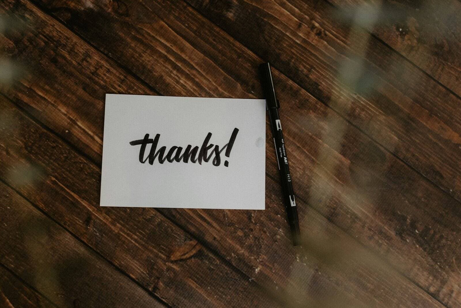 A white card lettered "thanks!" in black brush script beside a pen on a dark wooden table