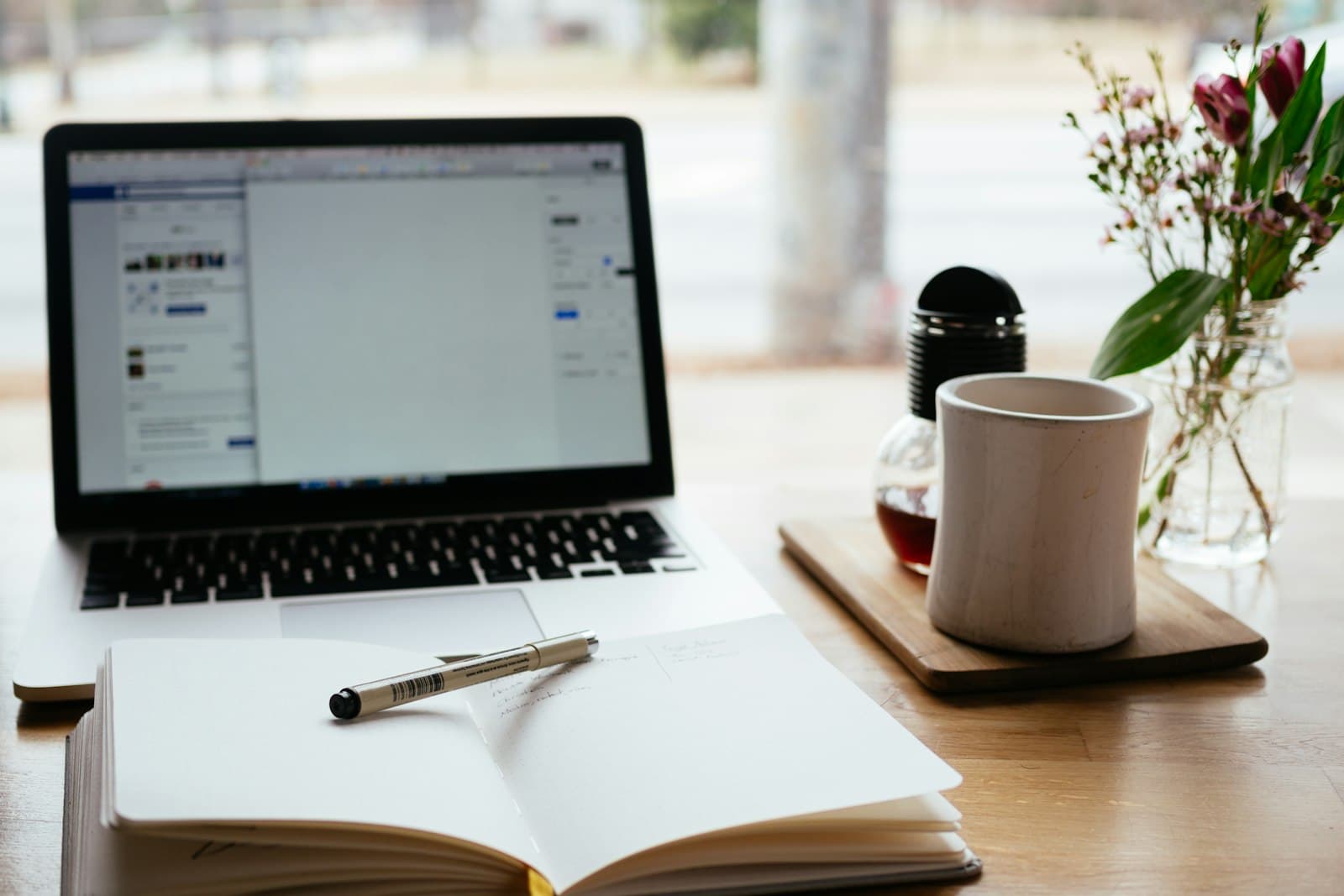 An open notebook and pen in front of a laptop, a mug and a small vase of flowers on a wooden table
