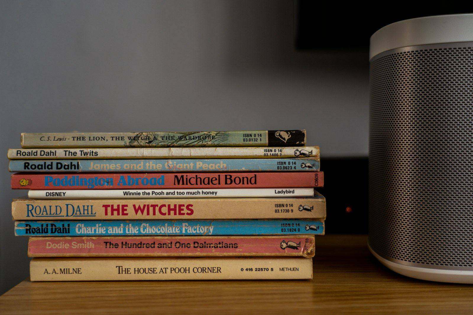 A stack of paperback books on a wooden shelf beside a speaker