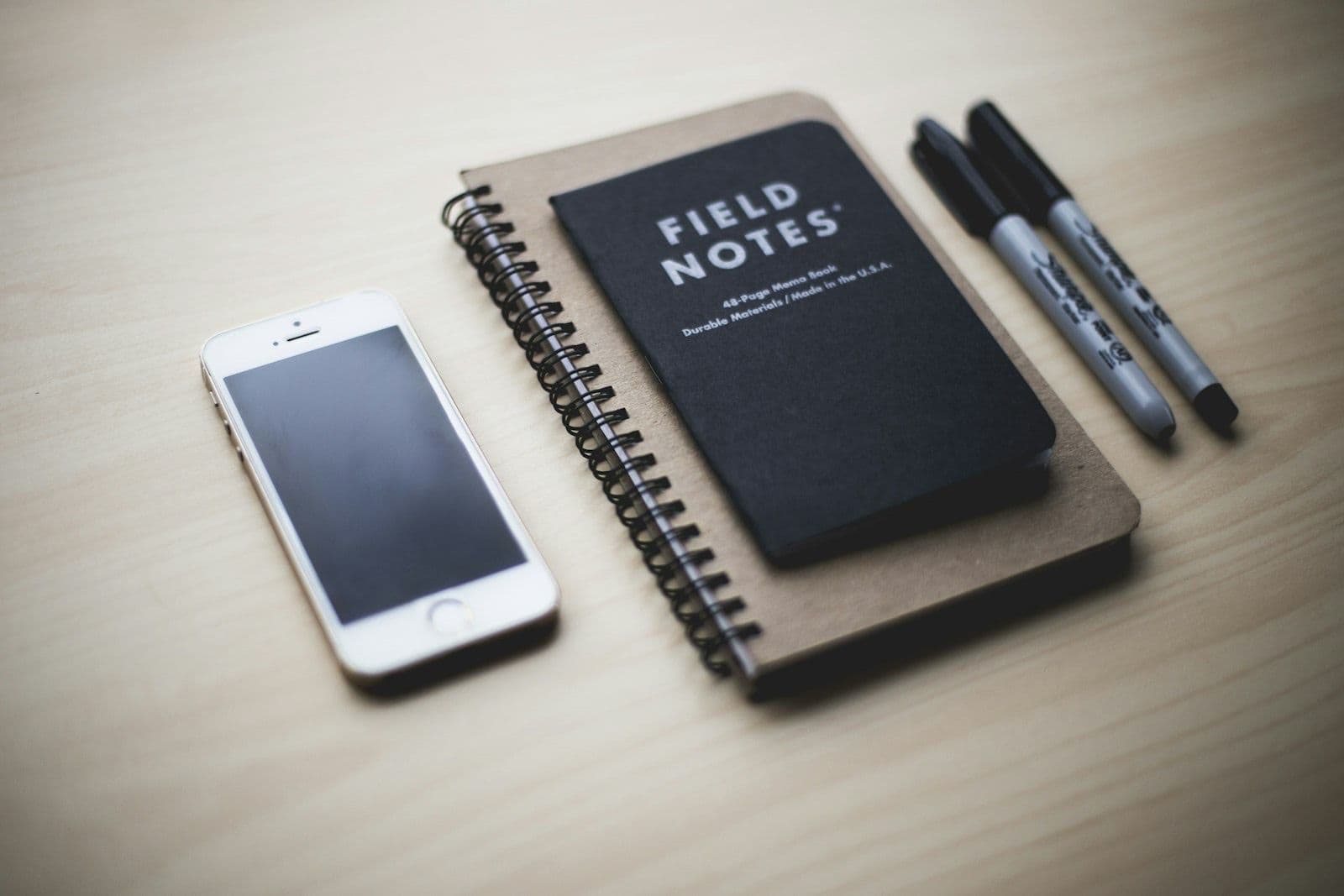 A phone resting beside a pocket notebook and pen on a pale wooden desk