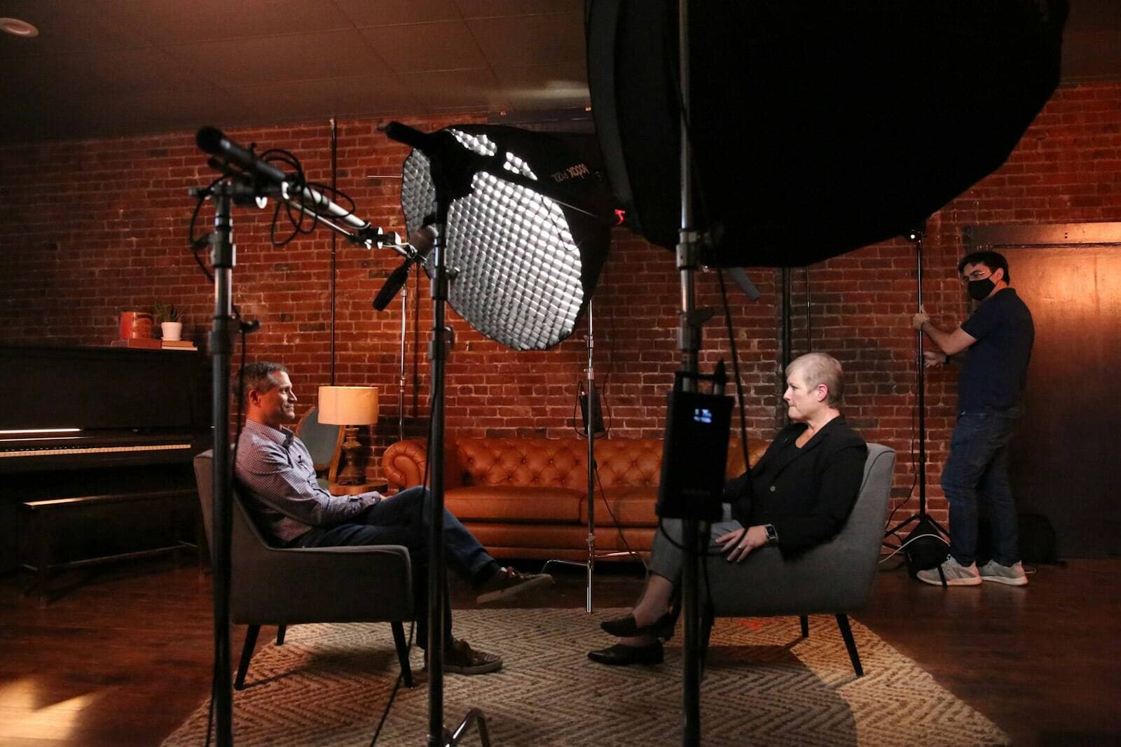 A man and a woman seated in chairs facing each other in a filming studio, waiting to record