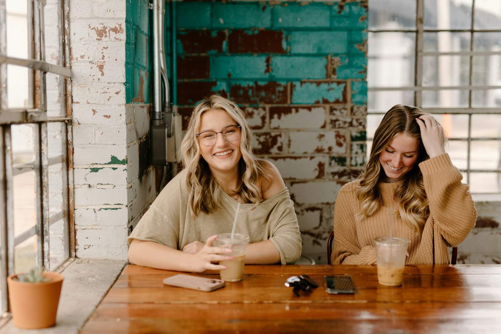 Two friends laughing side by side at a wooden table in front of a brick wall