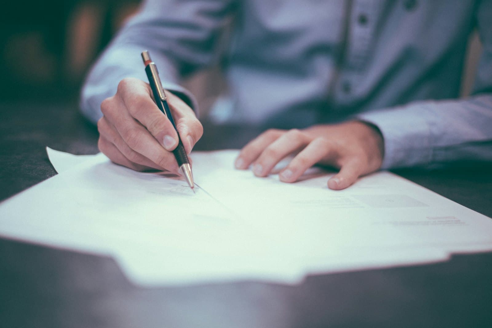 A person in a light blue shirt signing a sheet of paper with a pen at a dark wooden desk